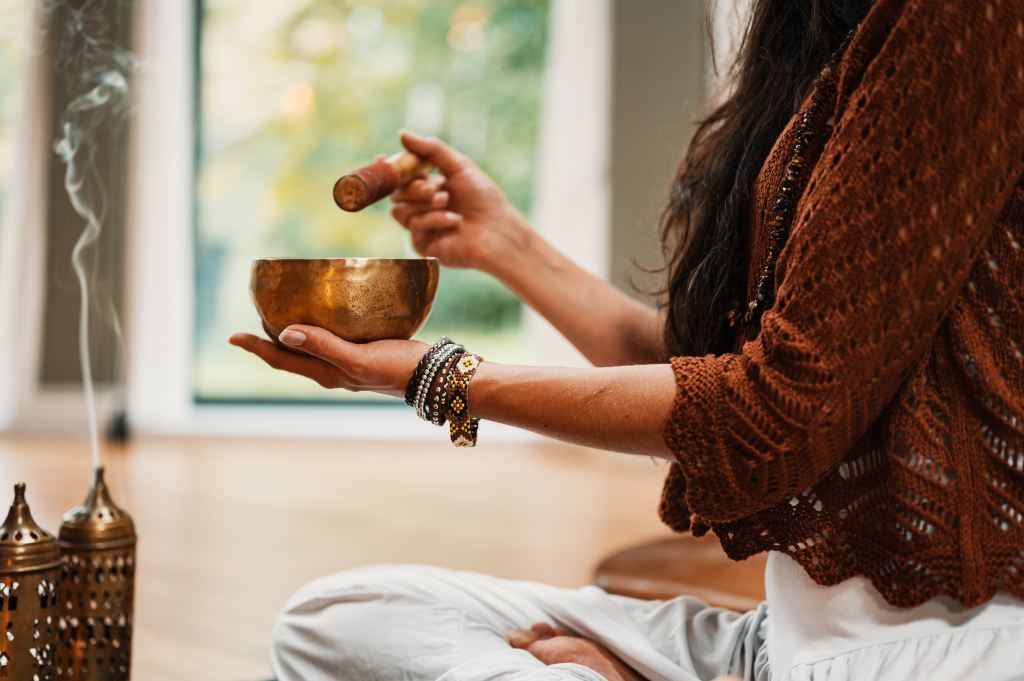 A close-up of a person gently striking a brass singing bowl with a mallet, while seated in a serene environment, with smoke rising in the background.